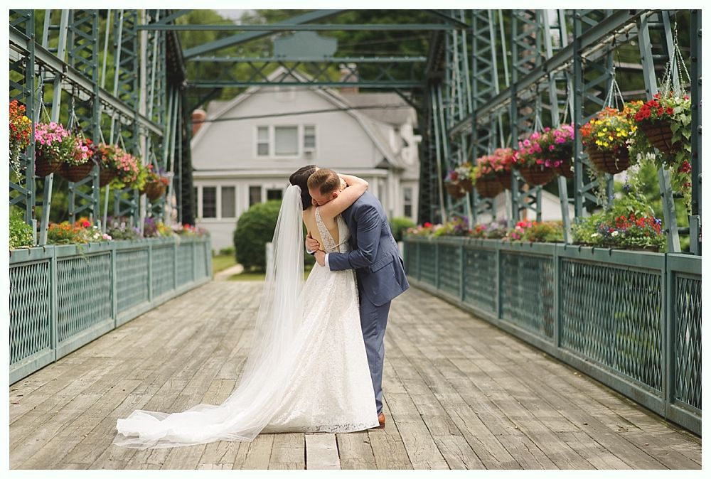 Bride and groom embrace on wooden bridge lined with flowers, house in background.