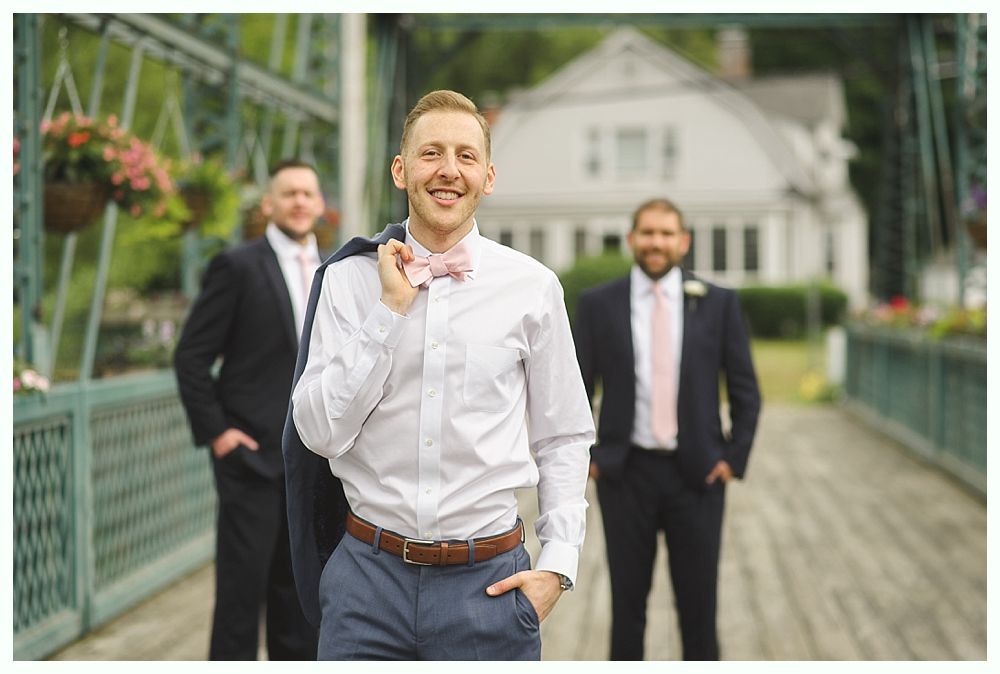 Groom and groomsmen on bridge, wearing suits and pink ties. Groom smiling, holding jacket. Building in background.