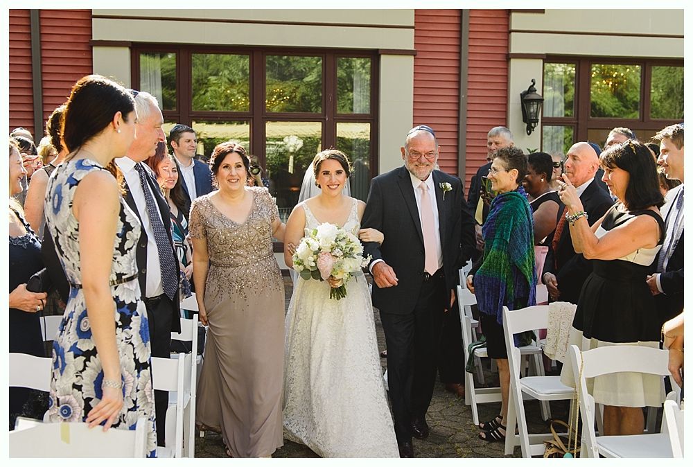 Bride escorted down aisle by two people, guests watching.