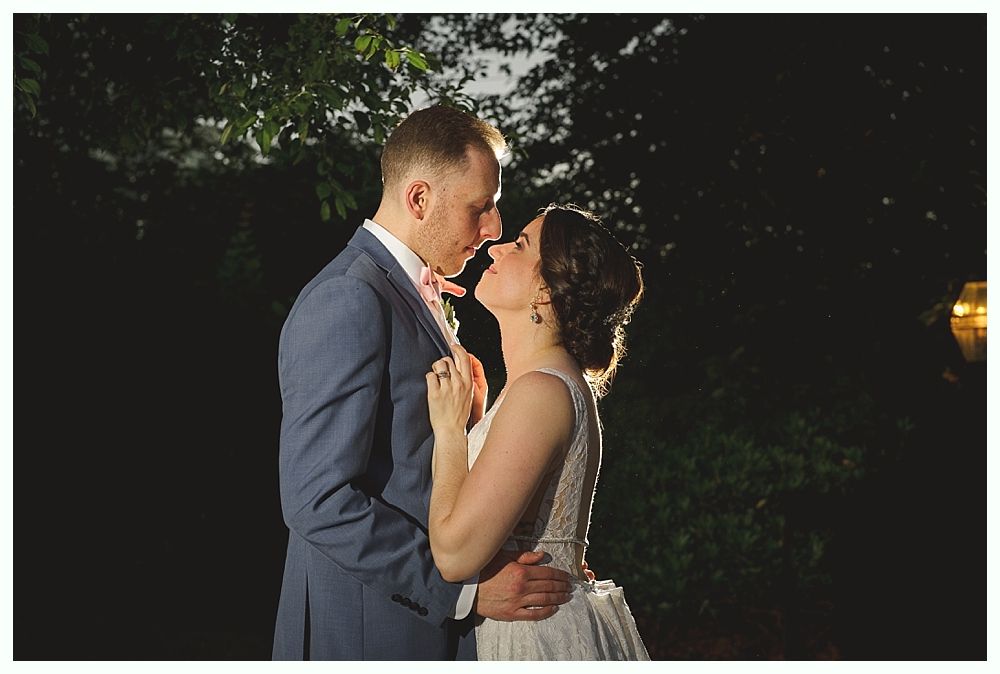 Bride and groom embrace outdoors at night, lit from behind.