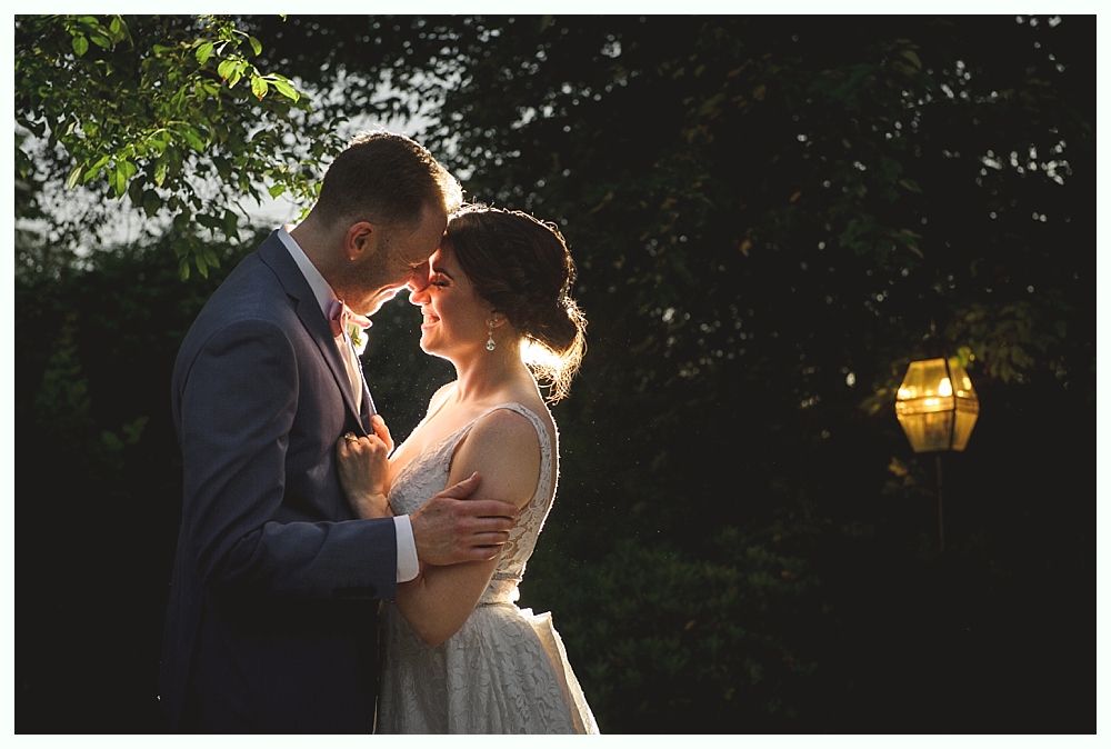 Couple embraces, backlit by sun, in a garden. The woman wears a white dress and the man a blue suit.