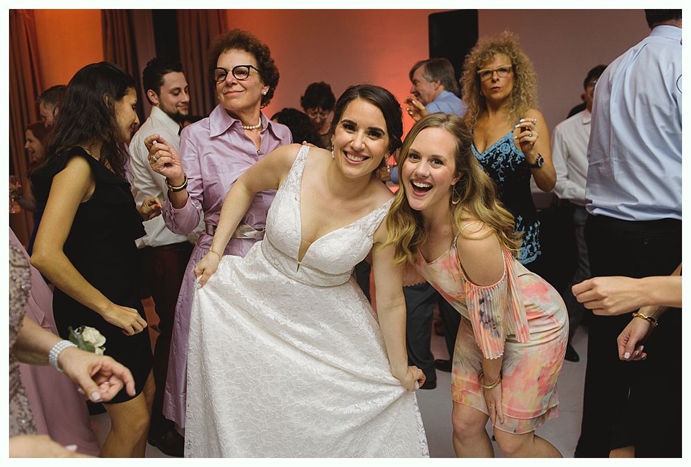 Bride dances with a friend, both smiling, surrounded by other wedding guests. Party setting.