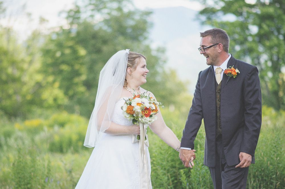 Bride walks down a path with two men, holding a yellow bouquet. Fountain and building in the background.