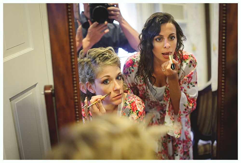 Two women applying makeup in front of a mirror, a photographer behind them, floral robes.