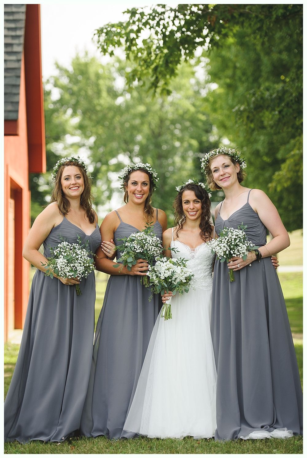 Bride and bridesmaids in a grassy field. Bride in white dress, bridesmaids in gray gowns, all holding bouquets.