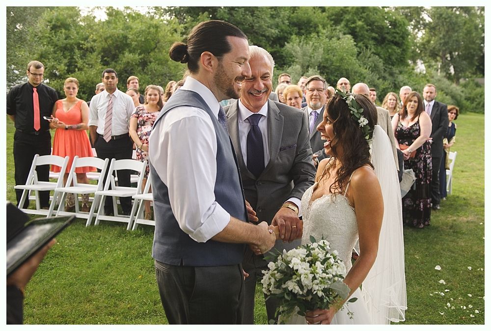 Wedding ceremony outdoors, couple holding hands, smiling. Guests watching.