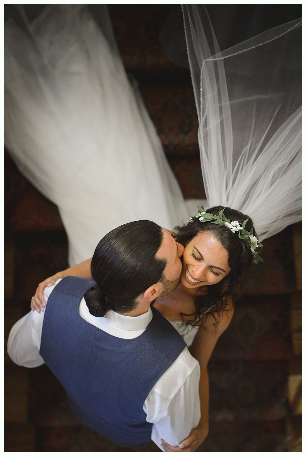 Groom kisses bride's cheek as they embrace; she smiles. Both wear wedding attire, under a flowing veil.