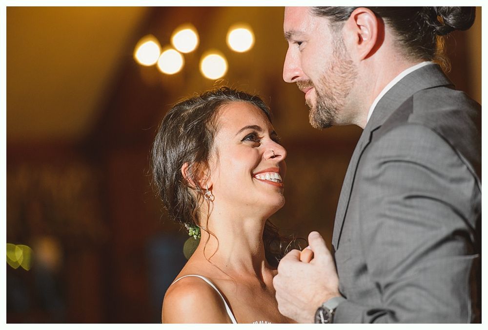 Bride and groom dance at reception, smiling at each other. Soft lighting, wooden interior.