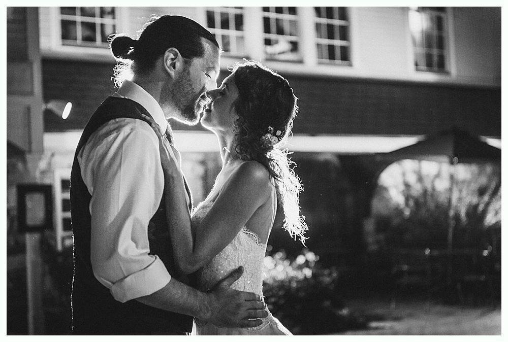 Newlyweds kiss tenderly outdoors at night, illuminated by soft light.