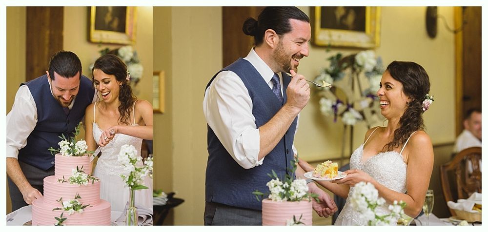 A wedding couple cuts cake, laughs, and feeds each other. Pink cake, white flowers, and a formal indoor setting.