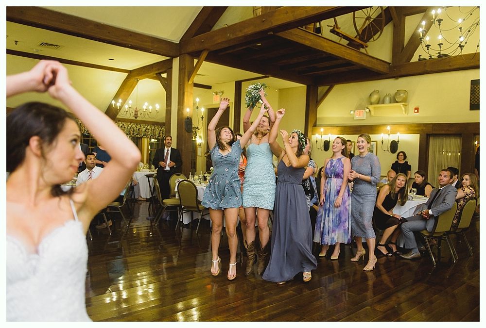 A wedding couple cuts cake, laughs, and feeds each other. Pink cake, white flowers, and a formal indoor setting.