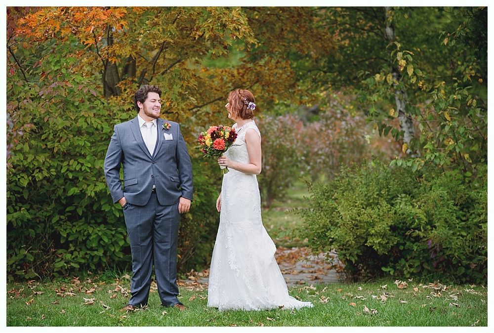 Couple in wedding attire facing each other outdoors, autumn foliage in the background.