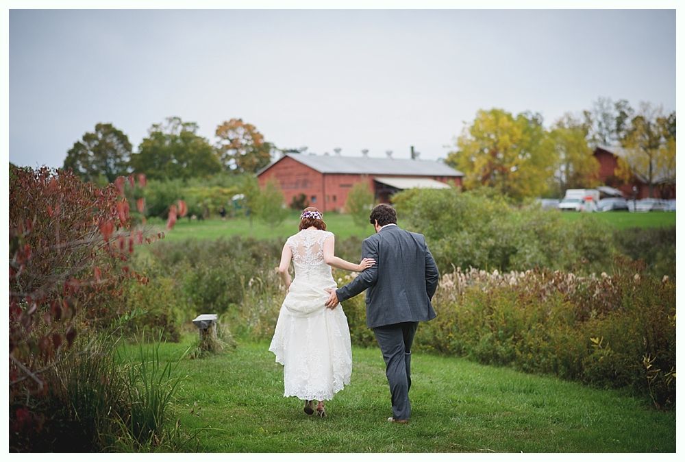 Bride and groom walking hand-in-hand away from camera, along a grassy path toward a red barn.