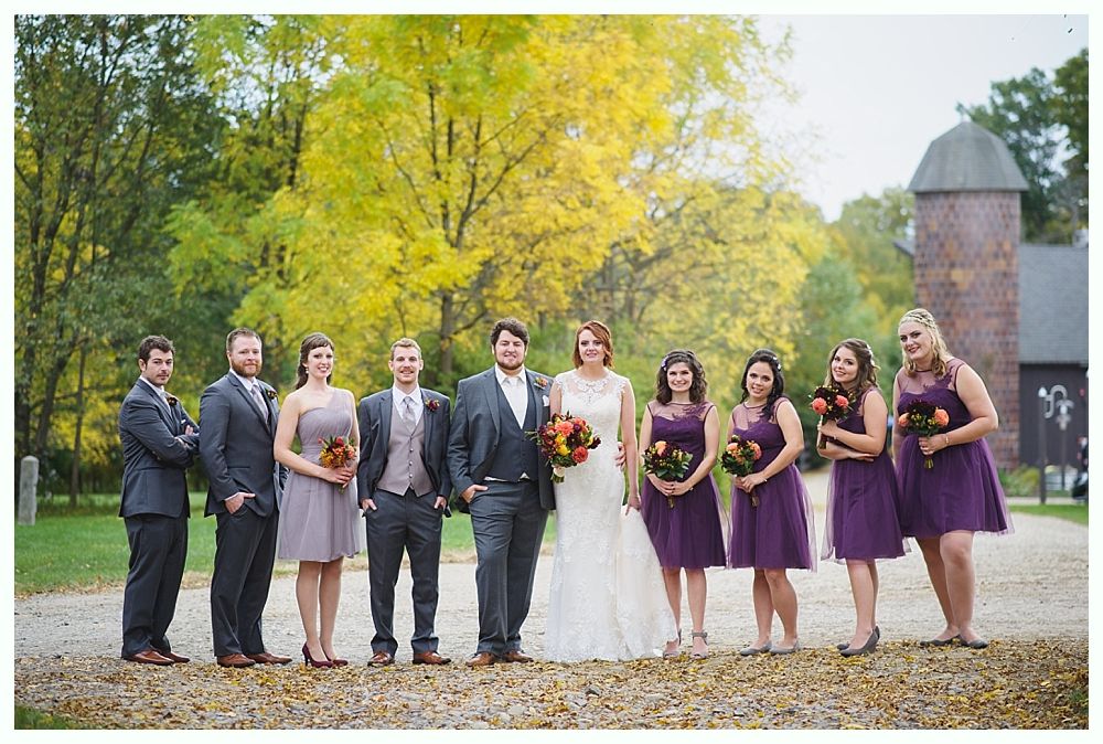 Wedding party in autumn: bride and groom with bridesmaids and groomsmen posing outside near barn.
