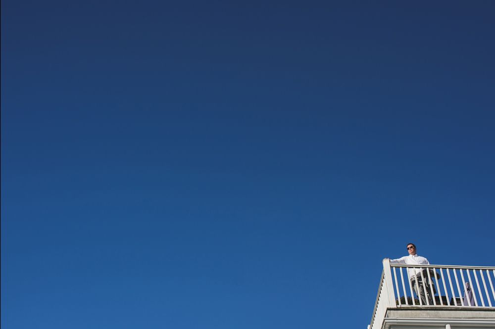 Person standing on a white balcony against a bright blue sky.