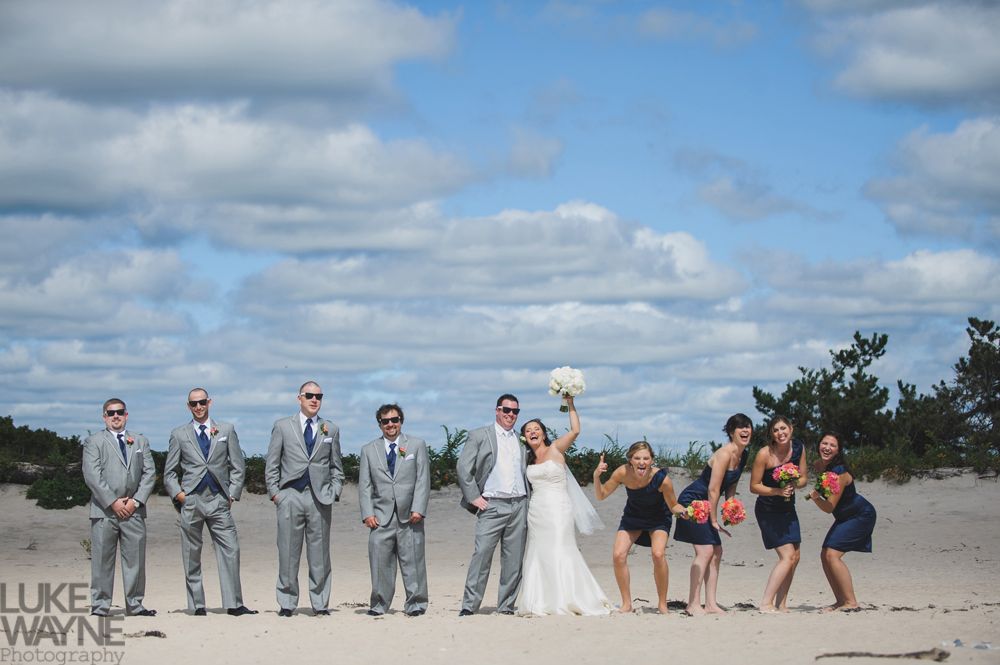 Wedding party poses on a beach, groom and bride with groomsmen and bridesmaids wearing suits and dresses, sunny sky.