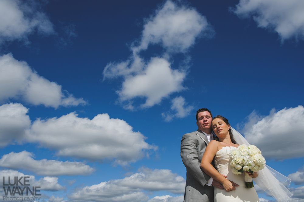 Bride and groom embrace outdoors against a bright blue sky with fluffy white clouds.
