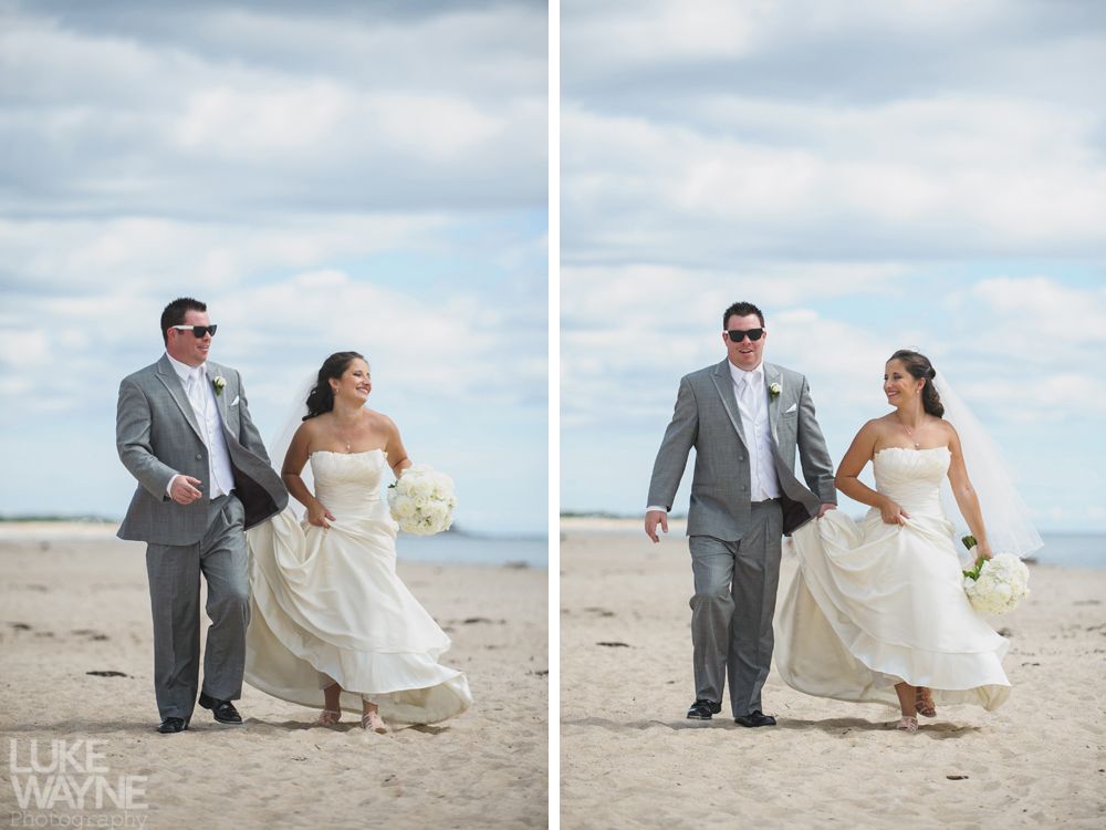 Bride and groom walking on a sandy beach, holding hands. Bride wears white dress and veil, groom in gray suit, both with sunglasses.