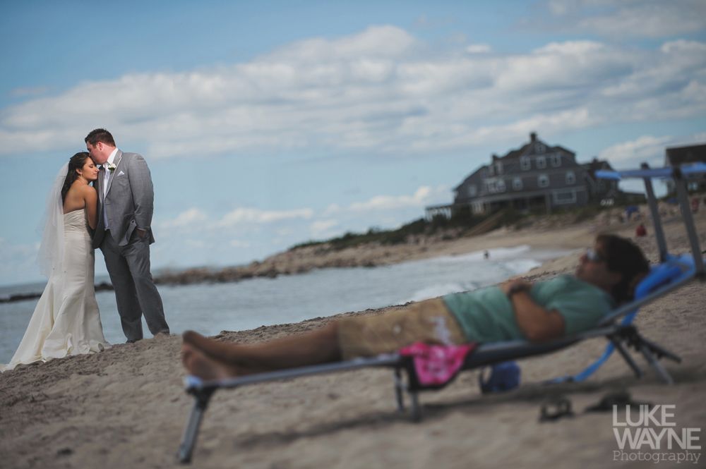 Bride and groom on beach, embracing. A person sunbathes in a chair nearby. Ocean and house in background.