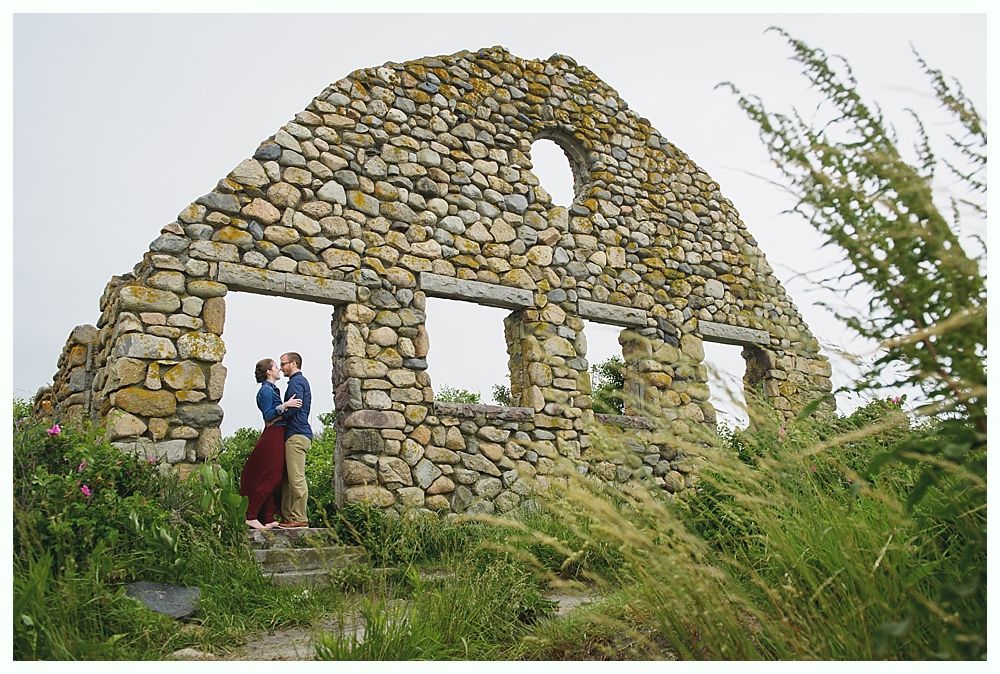 Couple kissing in front of stone ruins with arched roof and windows, surrounded by grass. Cloudy sky.
