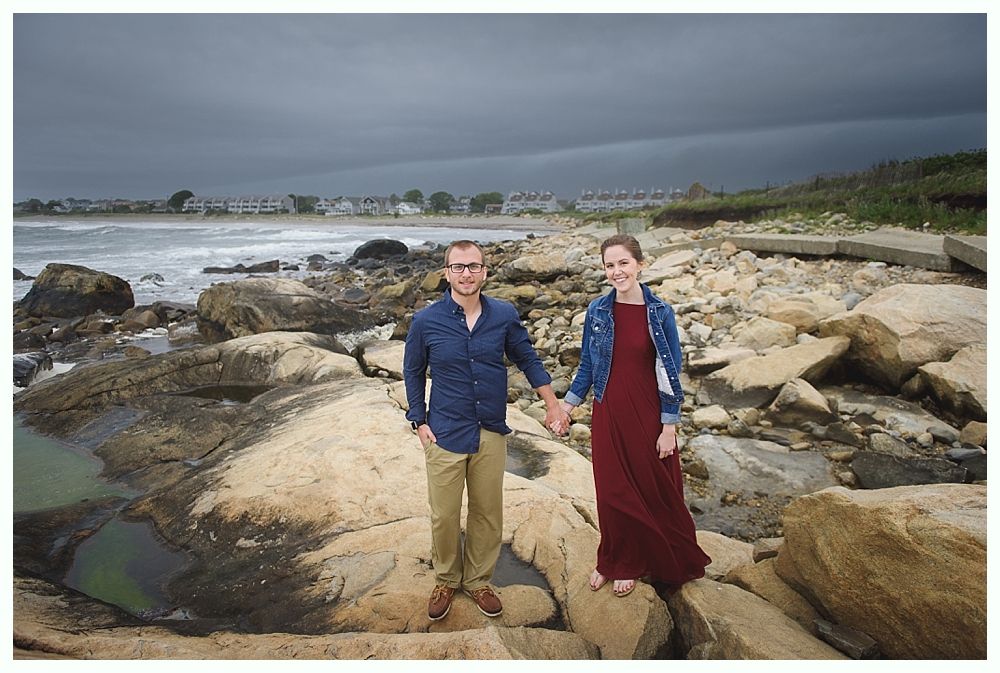 Couple holds hands on rocky beach; woman in red dress, man in blue shirt, cloudy sky.