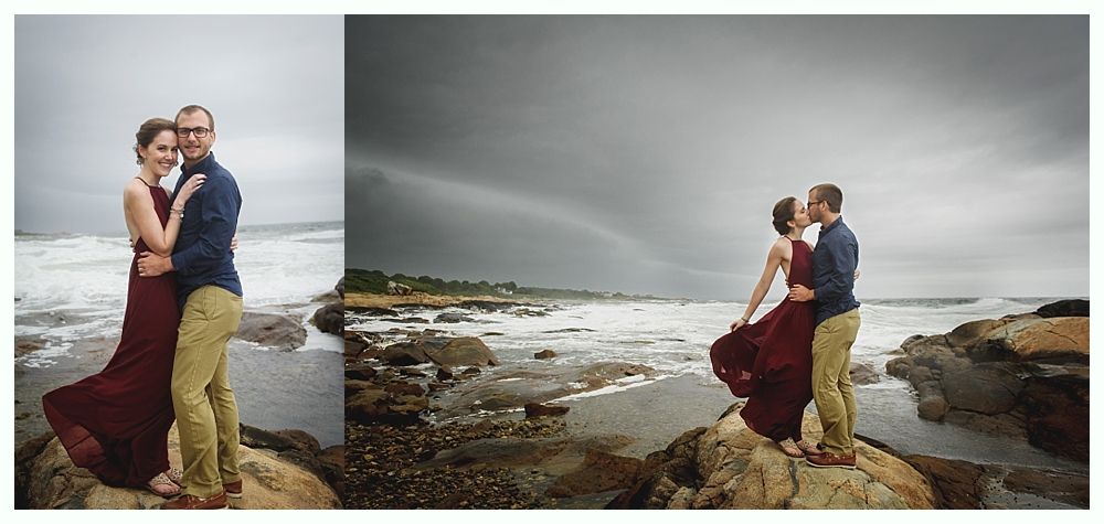 Couple kissing on rocky beach under a stormy sky with a rainbow. The woman wears a red dress, the man a blue shirt.