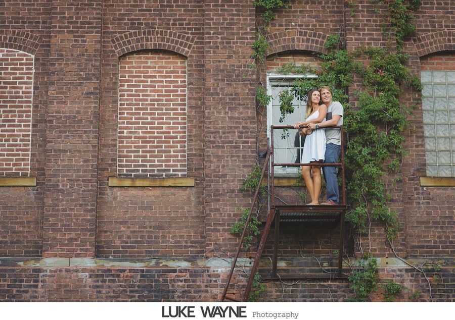 Couple standing on a rusty fire escape in front of a brick building.