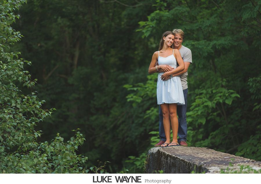 Couple embraces on stone structure, surrounded by green foliage. The woman wears a white dress; the man, a gray shirt.