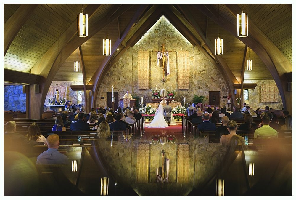 Wedding ceremony in a church with bride and groom at the altar, guests seated, and a large cross in the background.