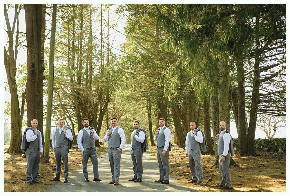 Group of men in gray suits stand on a tree-lined path, some holding suit jackets.