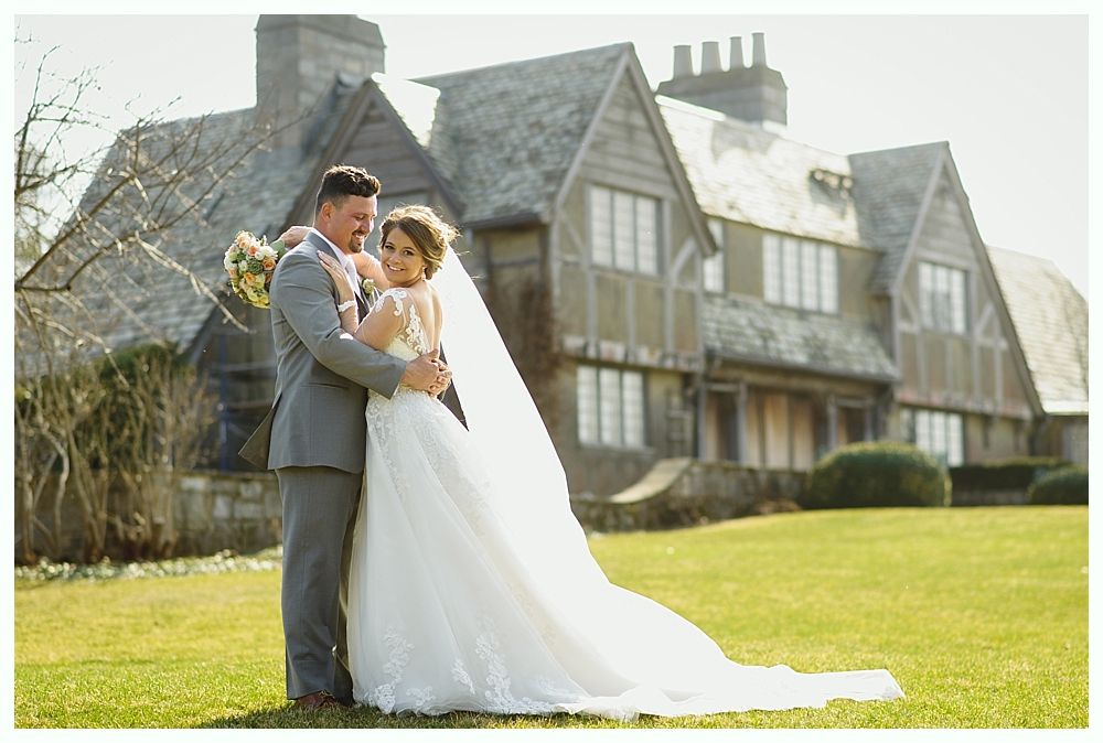Bride and groom embrace on a lawn in front of a large house. The bride wears a white gown with a long veil, and the groom wears a gray suit.