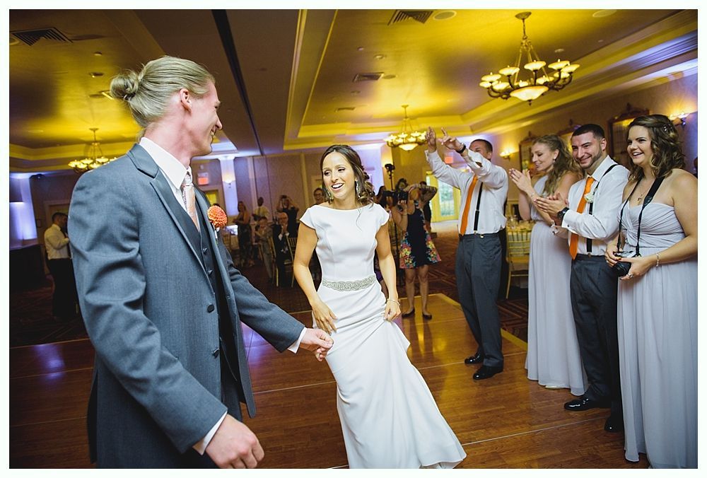 Bride with arms up, making a funny face, groom looking at his hand, cake cutting in background.