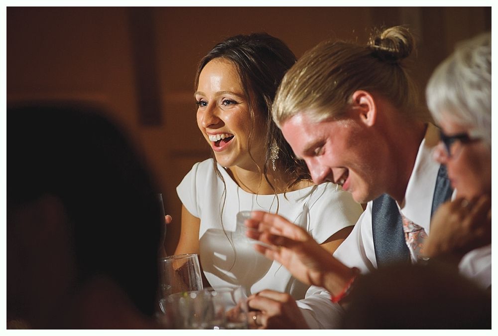Bride with arms up, making a funny face, groom looking at his hand, cake cutting in background.