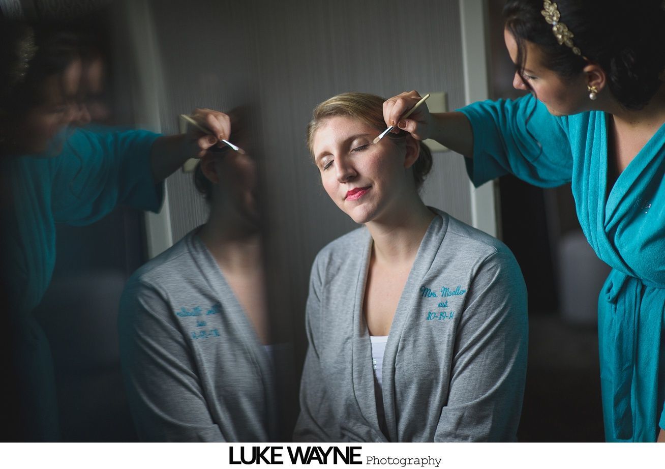 Woman having makeup applied, reflected in mirror. Person in teal robe applies makeup to woman in gray robe.