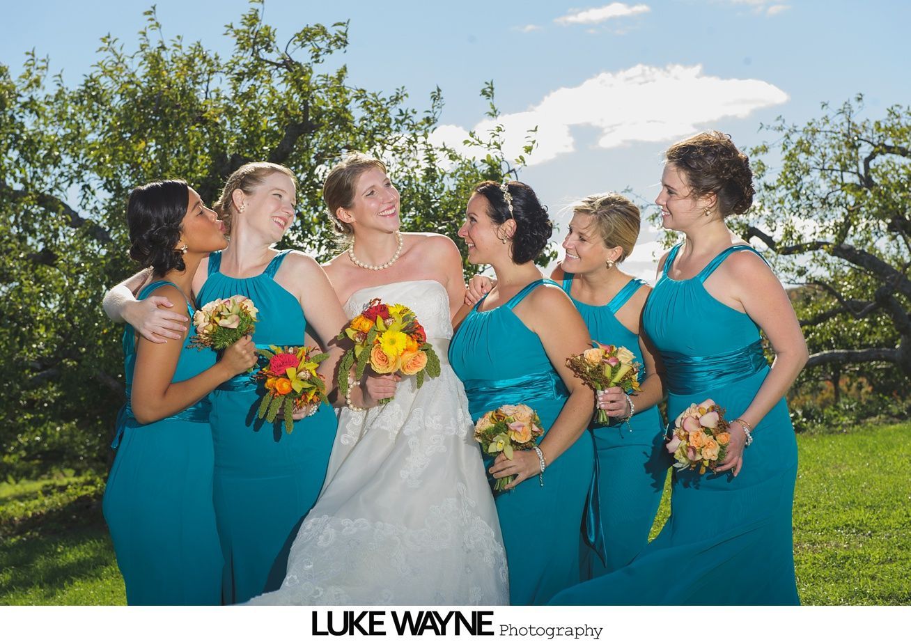 Bride smiles surrounded by teal-clad bridesmaids, holding bouquets in an orchard.