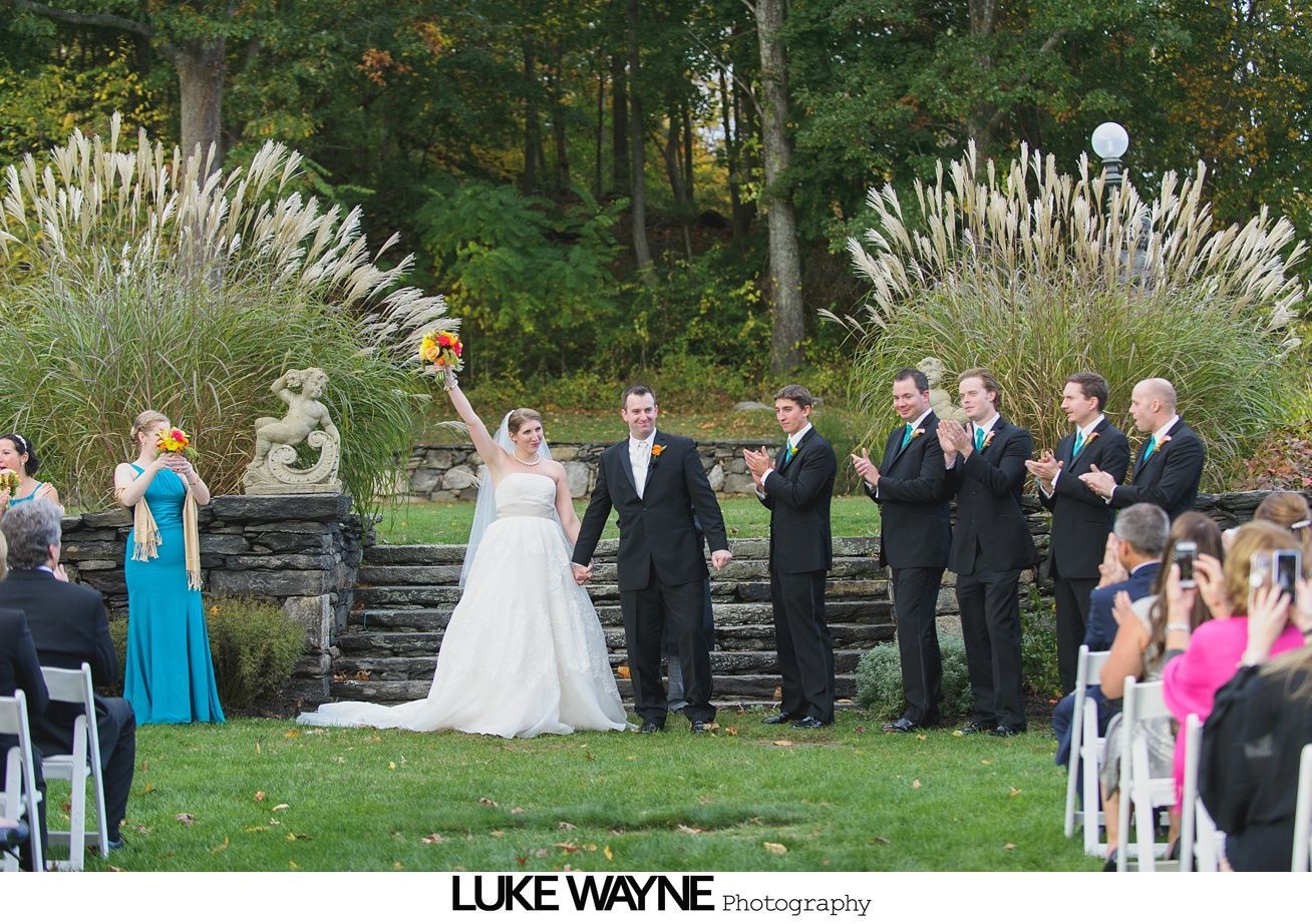 Bride and groom celebrating at an outdoor wedding ceremony, surrounded by the wedding party and guests.