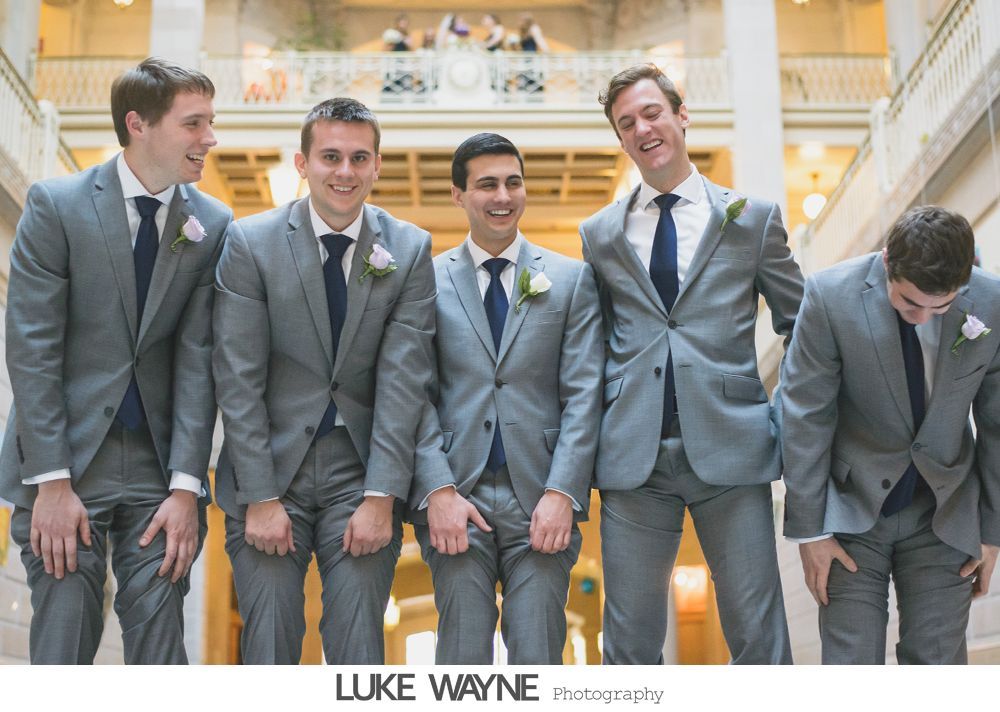 Six men in suits pose on steps by a street. One man has arms open, others smile or look at the camera. A bus and car are in the background.