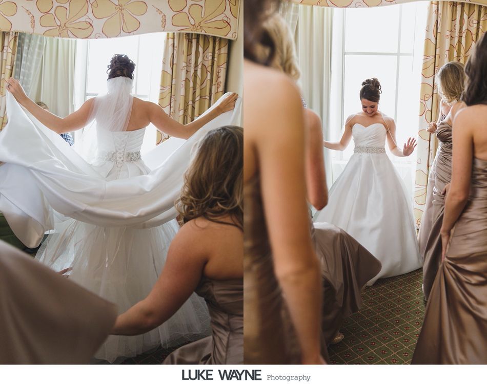 Bride in white gown, veil, arm raised, with bridesmaids in tan dresses. Hotel room with window.