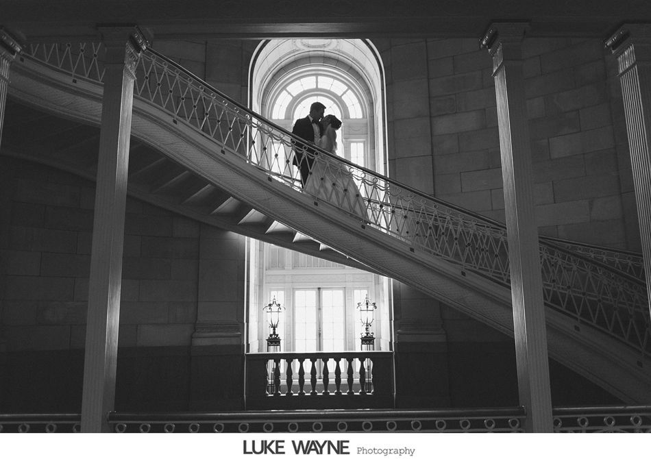 Bride and groom embracing on a grand staircase, framed by a large arched window. Black and white.
