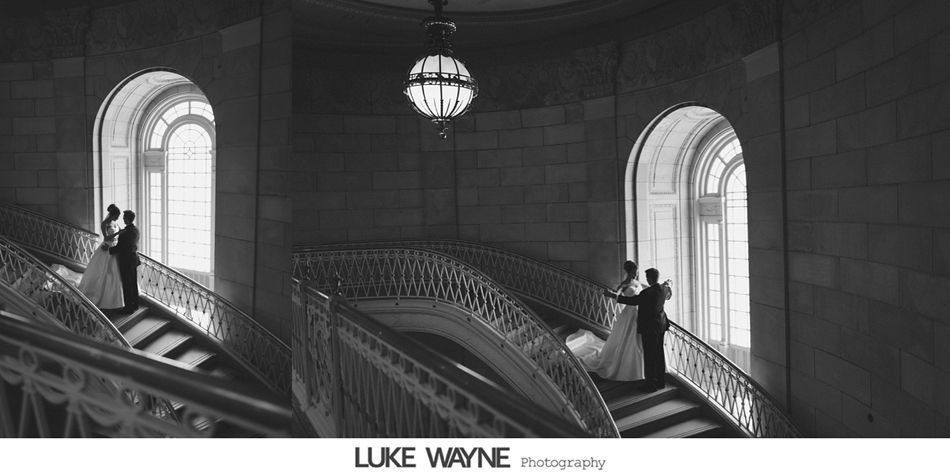 A wedding couple stands on a staircase, embracing near a large window, in a grand hall. Black and white.