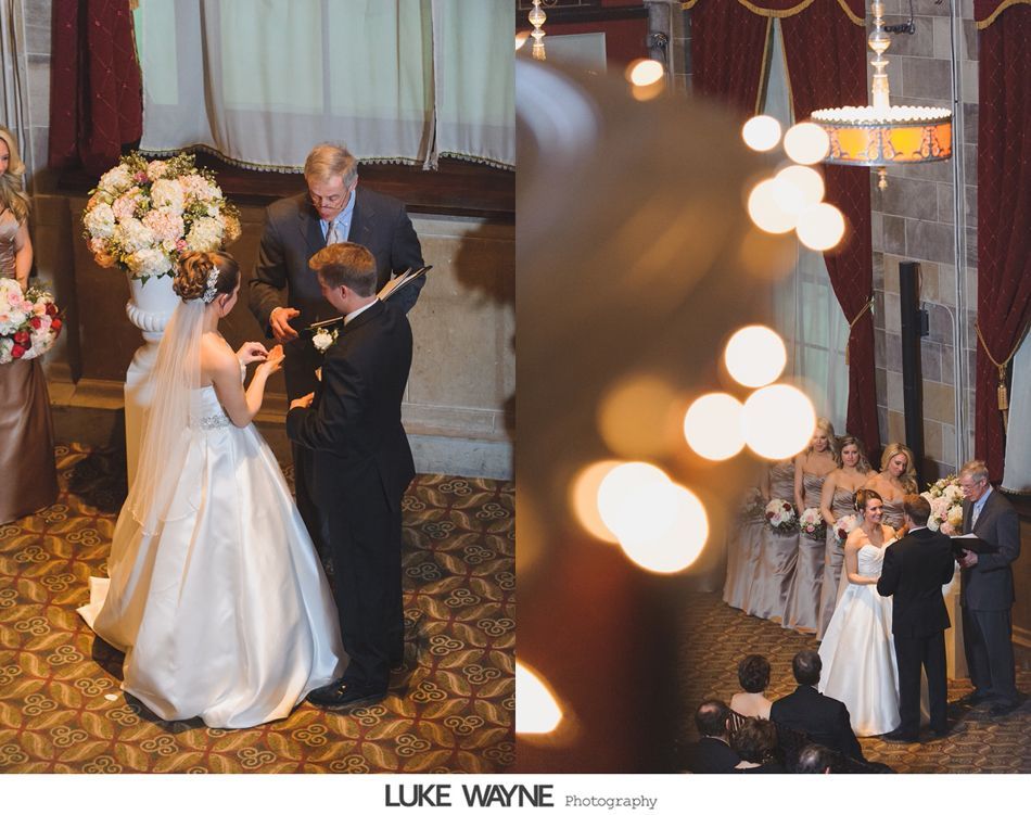 Wedding ceremony, couple exchanging rings, officiant, bridesmaids, floral arrangement, elegant interior with curtains and chandelier.