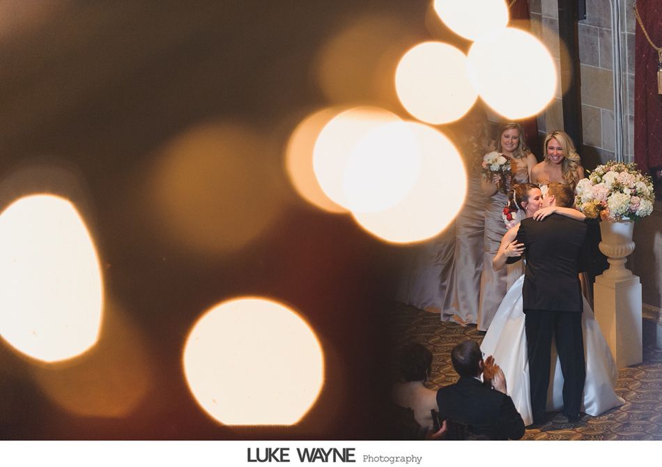 Wedding couple hugging; bridesmaids clap; warm lights in foreground.
