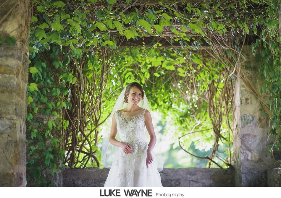 Bride in a white gown and veil smiles under a vine-covered stone arbor.