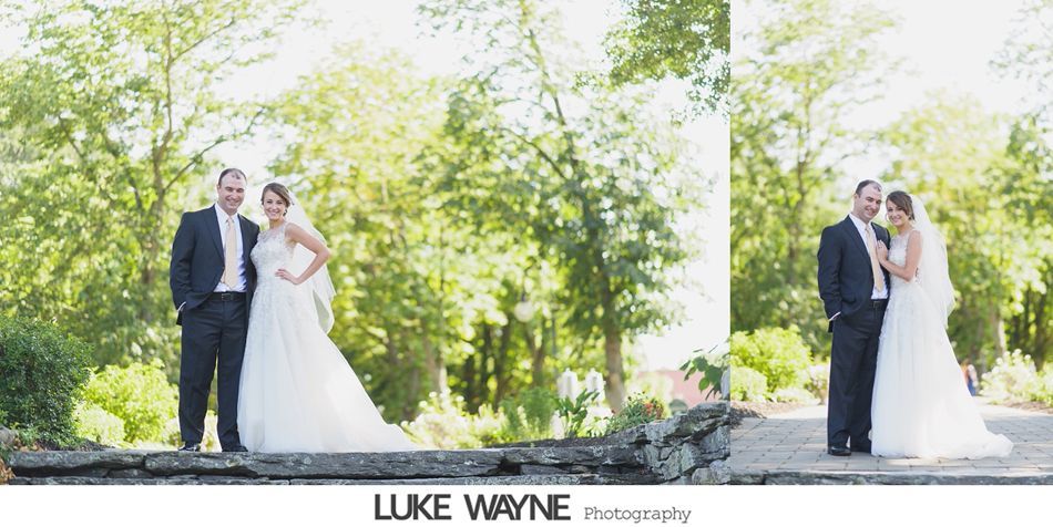 Bride and groom pose on a stone wall in a park. The bride wears a white dress and veil. The groom wears a suit.