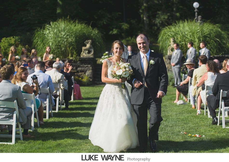 Bride and father walk down aisle at outdoor wedding ceremony; lush greenery surrounds.