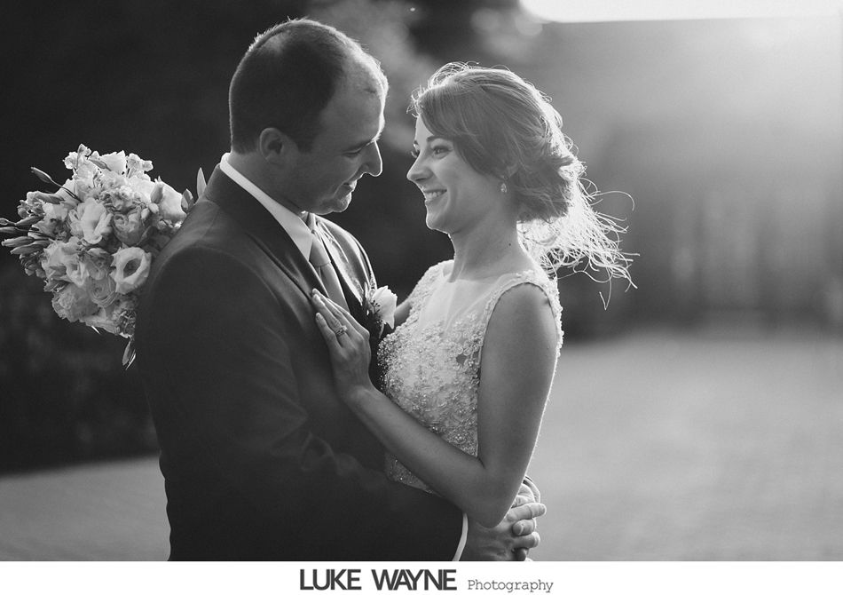 Bride and groom embrace, smiling. Bride in dress, groom in suit, holding bouquet; backlit.