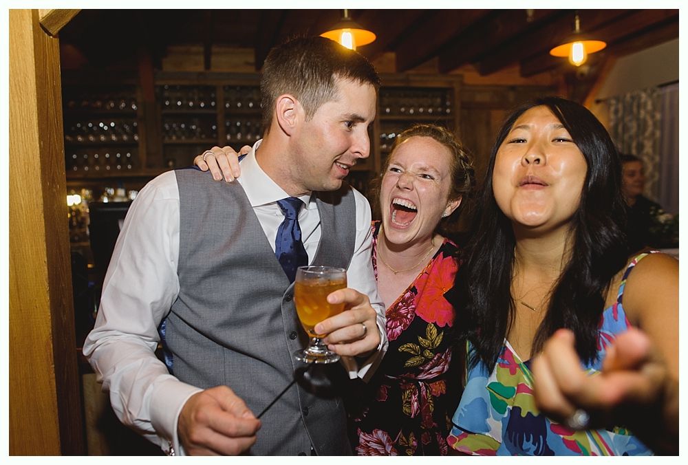Three people laughing, celebrating. Man in a vest holds a drink, woman next to him laughs widely, and another woman points and smiles.