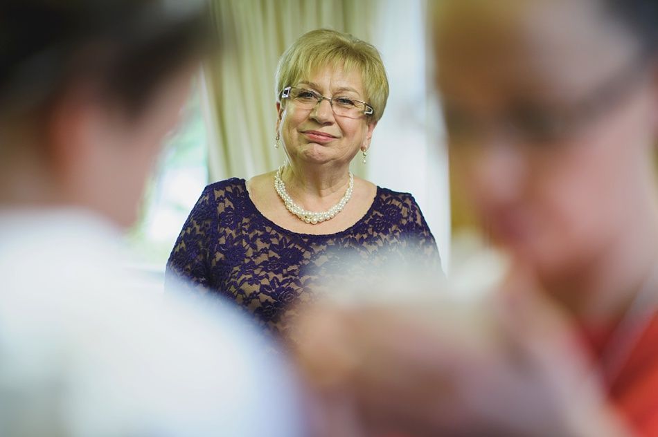 Woman in a dark purple lace dress and pearl necklace looks towards others in a bright room.