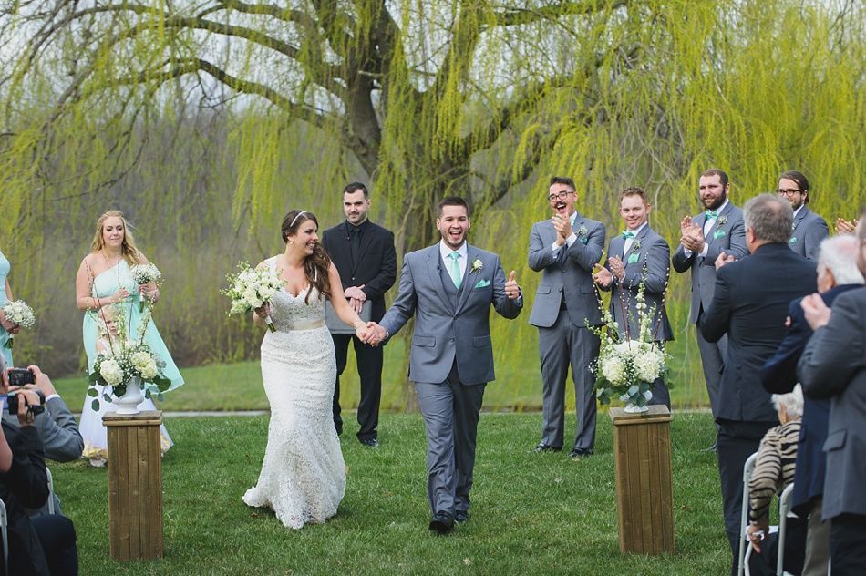 Newly married couple walks down the aisle, cheered by wedding party and guests, under a willow tree.
