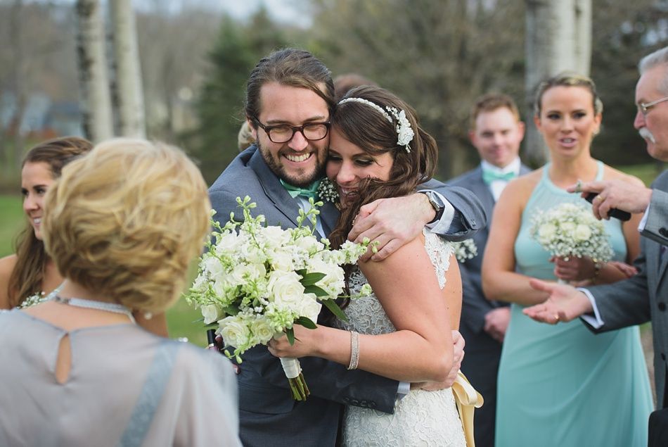 Bride and groom embrace, holding bouquet, surrounded by wedding guests. Outdoor ceremony.